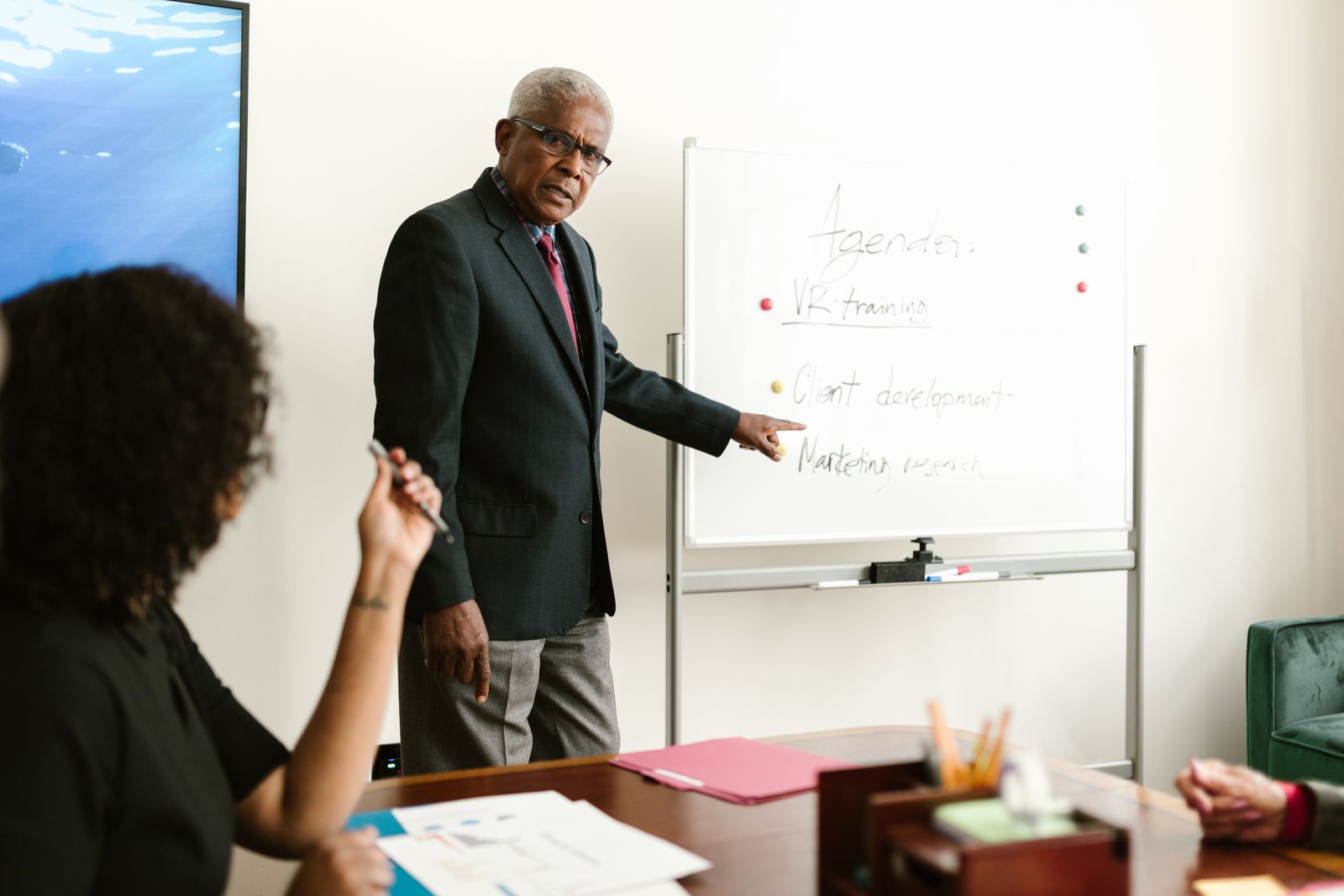 An elderly man in business attire leading a meeting and discussing plans on a whiteboard.